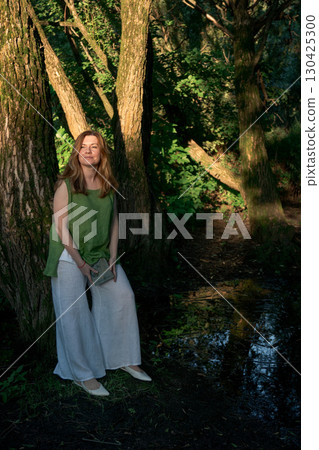 Woman in a green linen top and white trousers leans against a tree while holding an open book in Izmailovsky Park, Moscow Woman in a green linen top and white trousers leans against a tree while holding an open book in Izmailovsky Park, Moscow 130425300