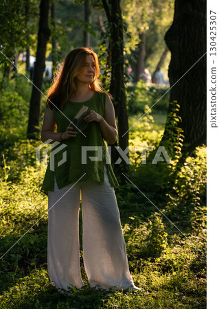 Woman in a green linen top and white trousers holds an open book in Izmailovsky Park, Moscow, standing in golden sunlight among lush greenery Woman in a green linen top and white trousers holds an open book in Izmailovsky Park, Moscow, standing in golden sunlight among lush greenery 130425307