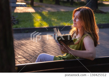 A woman sits on a park bench at sunset, reading an old book while warm golden light highlights her hair and face A woman sits on a park bench at sunset, reading an old book while warm golden light highlights her hair and face 130425312