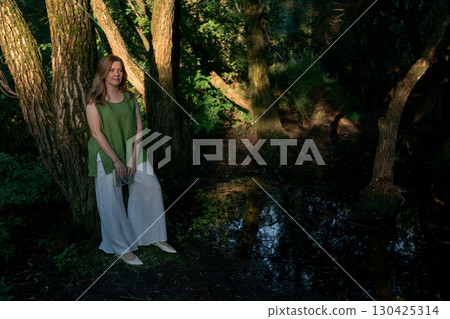 Woman in a green linen top and white trousers leans against a tree while holding an open book in Izmailovsky Park, Moscow Woman in a green linen top and white trousers leans against a tree while holding an open book in Izmailovsky Park, Moscow 130425314