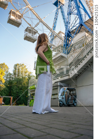 A woman in a green linen top and white trousers stands near a Ferris wheel in Izmailovsky Park, gazing upward against the clear sky A woman in a green linen top and white trousers stands near a Ferris wheel in Izmailovsky Park, gazing upward against the clear sky 130425315