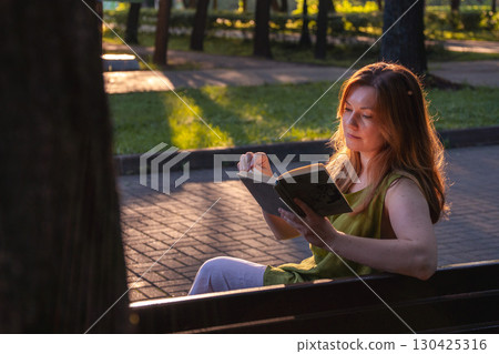A woman sits on a park bench at sunset, reading an old book while warm golden light highlights her hair and face A woman sits on a park bench at sunset, reading an old book while warm golden light highlights her hair and face 130425316
