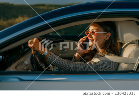A Stylish Woman is Driving a Car while Engaged in a Phone Call during a Beautiful Day 130425335