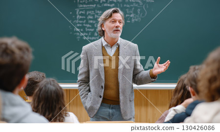 Mature male professor giving a lecture in classroom while standing in front of a chalkboard with handwritten notes 130426086