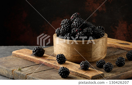 A wooden bowl filled with ripe blackberries on a board. A few berries are scattered around 130426207