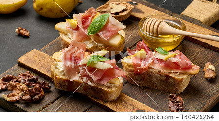 Italian bruschetta with prosciutto, pear slices, cream cheese and fresh basil, garnished with sesame seeds, served on a rustic wooden board 130426264