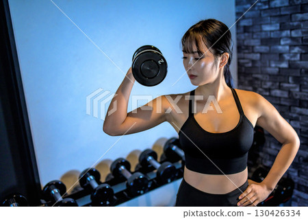 A woman lifting a dumbbell with one hand. A cool woman exercising at the gym. Upper body. Muscle training girl. A woman lifting a dumbbell with one hand. A cool woman exercising at the gym. Upper body. Muscle training girl. 130426334