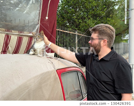 A man petting a cat resting on the roof of a red car. Human-animal connection, affection, and companionship. 130426391