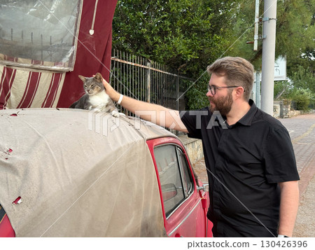 A man petting a cat resting on the roof of a red car. Human-animal connection, affection, and companionship. A man petting a cat resting on the roof of a red car. Human-animal connection, affection, and companionship. 130426396