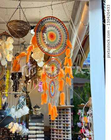 Colorful dreamcatchers with orange feathers hanging at a souvenir shop. Tradition, spirituality and tourism represented through handmade crafts, decoration and cultural items. Colorful dreamcatchers with orange feathers hanging at a souvenir shop. Tradition, spirituality and tourism represented through handmade crafts, decoration and cultural items. 130426433