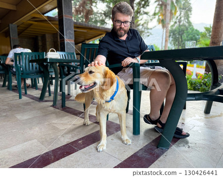 Man petting a golden dog with blue collar at outdoor table. Friendship, companionship and leisure represented through human animal bond, touch and interaction. 130426441