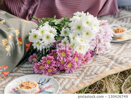 Bouquet of white and purple chrysanthemums lies on a patterned blanket during a picnic. Nearby, ceramic plates and a person with floral fabric create a cozy setting in the grass Bouquet of white and purple chrysanthemums lies on a patterned blanket during a picnic. Nearby, ceramic plates and a person with floral fabric create a cozy setting in the grass 130426749