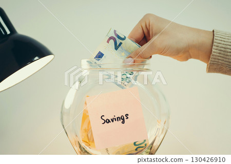 Collecting savings from income concept, toned image. Person putting 20 euro banknote into the glass jar Collecting savings from income concept, toned image. Person putting 20 euro banknote into the glass jar 130426910