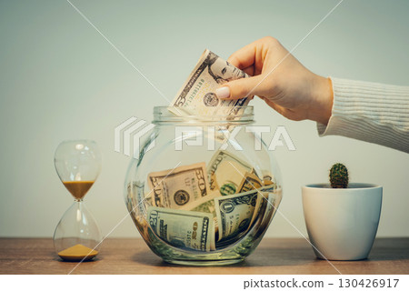 Female hand putting dollars money into glass jar, toned. Saving money, leaving part of salary to save for some big purchase Female hand putting dollars money into glass jar, toned. Saving money, leaving part of salary to save for some big purchase 130426917
