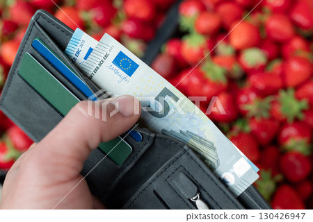 Wallet with euro money and credit cards in hand of a man, fresh strawberries on the background, close up. Concept of organic farming, earning money in the countryside, seasonal purchase 130426947