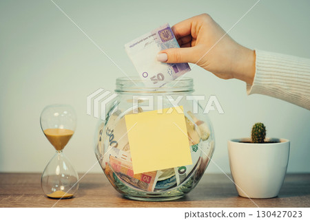 Ukrainian person saving up money for a big purchase. Girl puts 50 hryvnias banknote to the glass jar with blank paper for inscription, toned photo Ukrainian person saving up money for a big purchase. Girl puts 50 hryvnias banknote to the glass jar with blank paper for inscription, toned photo 130427023