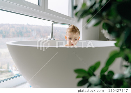 The boy enjoys a relaxing bath in the spacious white bathtub , the large bathroom is flooded with sunlight thanks to large floor-to-ceiling windows. 130427547