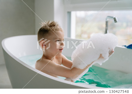 Bathed in natural light, the boy leans back in the white tub filled with calm, clear water, while the wide windows frame a peaceful view beyond. High quality photo 130427548