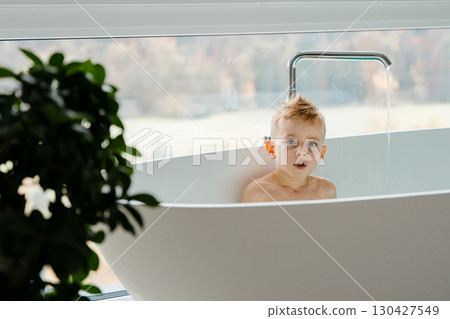 The boy, around five years old, laughs as he creates little waves with his toy boat, the spacious white tub around him shimmering with turquoise water under the daylight. High quality photo 130427549