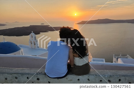 Romantic sunset in Santorini. A couple sits closely on a stone wall overlooking the sea at sunset in Santorini, Greece, with the islands iconic white church and blue dome in view 130427561