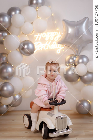 Baby girl in pink hoodie riding toy car on wooden floor with balloons and birthday sign Baby girl in pink hoodie riding toy car on wooden floor with balloons and birthday sign 130427759