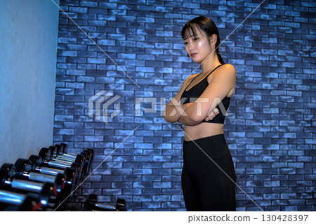 一位女士正在選擇啞鈴的重量。一位酷酷的女士正在健身房鍛鍊。一位女士正在進行肌肉訓練。 130428397