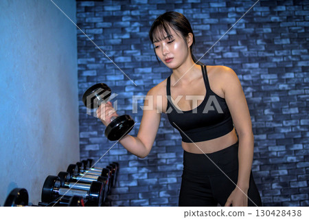一位女士正在選擇啞鈴的重量。一位酷酷的女士正在健身房鍛鍊。一位女士正在進行肌肉訓練。 130428438