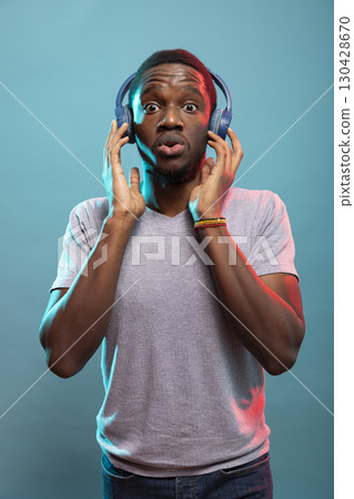Young black man in headphones standing against blue background, expressing surprise with wide eyes and pursed lips. Shocked african american person touching his wireless headset and looking at camera. 130428670