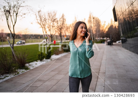 Happy young woman walking and talking on the phone near office building 130428693