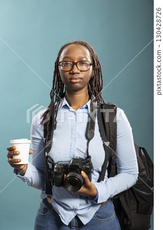 Female photographer standing with camera and disposable cup in her hands. African American female model poses confidently, wearing blue shirt, with DSLR around neck and holding coffee. Female photographer standing with camera and disposable cup in her hands. African American female model poses confidently, wearing blue shirt, with DSLR around neck and holding coffee. 130428726