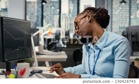 Equity analyst in office reviews quarterly earnings paperwork to assess future stock market value, talking on phone. Trader making calls, conducting financial analysis to improve investments 130428743