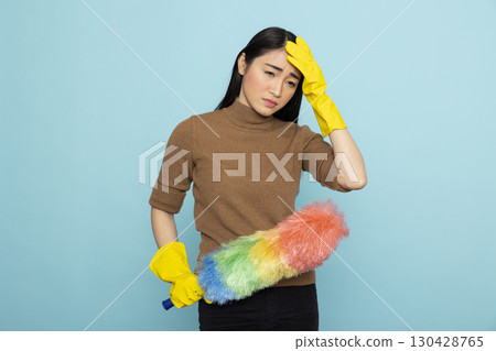 Portrait of overwhelmed cleaning service staff holding feather duster, experiencing job fatigue. Asian housemaid in yellow rubber gloves expressing exhaustion, standing isolated over blue background. 130428765