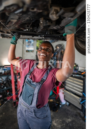 Young female african mechanic working under a car in a garage 130428777