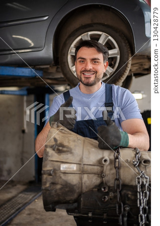 Mechanic holding a gearbox and showing thumbs up in a workshop 130428779
