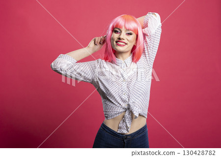 Beautiful female individual standing in relaxed stretching posture, smiling confidently at camera. Cheerful young woman in casual clothing, posing with arms behind head against pink background. 130428782