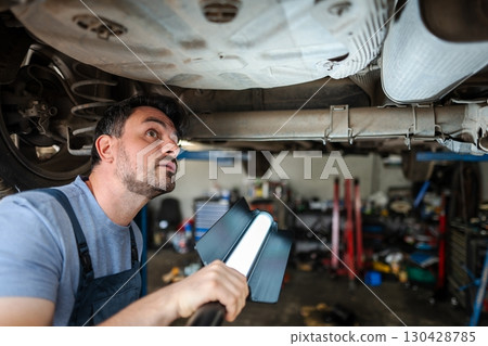 Mechanic inspecting car chassis with a lamp in a garage Mechanic inspecting car chassis with a lamp in a garage 130428785