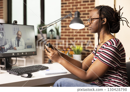 African american businesswoman holding medicine box while discussing treatment with physician on telehealth videocall. Black female freelancer holding pills bottle, talking with male doctor remotely. 130428798