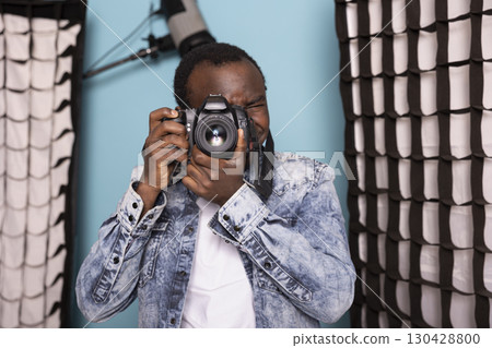 Black male photographer, deeply focused, using his DSLR camera in a studio setting. African american man positioned near softboxes and professional lighting, preparing to capture high quality images. Black male photographer, deeply focused, using his DSLR camera in a studio setting. African american man positioned near softboxes and professional lighting, preparing to capture high quality images. 130428800