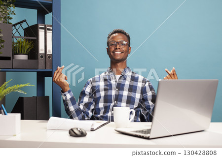 Smiling young man gesturing for cash by rubbing his fingers in a playful payday moment on a blue background. Cheerful black guy seated at his desk is expressing excitement over money rewards. Smiling young man gesturing for cash by rubbing his fingers in a playful payday moment on a blue background. Cheerful black guy seated at his desk is expressing excitement over money rewards. 130428808