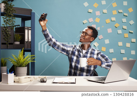 Young black accountant with coffee in hand, seated at office desk, taking a selfie with his mobile device. African american manager holding a cup and cellphone, recording his workday. 130428816