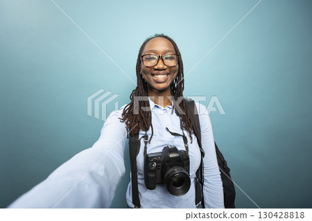 Pov of african american woman holding camera to record herself, grinning as she creates content in studio with dslr around her neck. Black female photographer smiling and taking a self portrait. 130428818