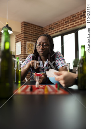 Skeptical african american woman with unsure expression, looking through cards, deciding on next move during board game session. The moment captures her deep thought as she considers options. Skeptical african american woman with unsure expression, looking through cards, deciding on next move during board game session. The moment captures her deep thought as she considers options. 130428834