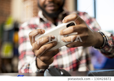 Hands of male entrepreneur holding cellphone, takes break from remote work, playing mobile games at desk. Young freelancer enjoying digital entertainment in cozy apartment home office. Hands of male entrepreneur holding cellphone, takes break from remote work, playing mobile games at desk. Young freelancer enjoying digital entertainment in cozy apartment home office. 130428872