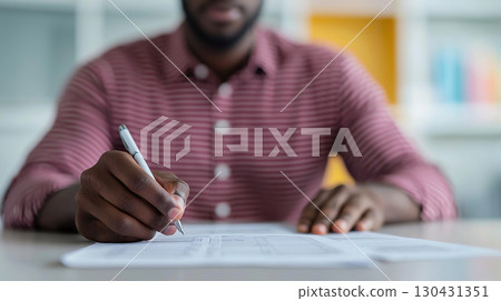 person in striped shirt is focused on filling out paperwork at desk, holding pen. background is softly blurred, suggesting office environment 130431351