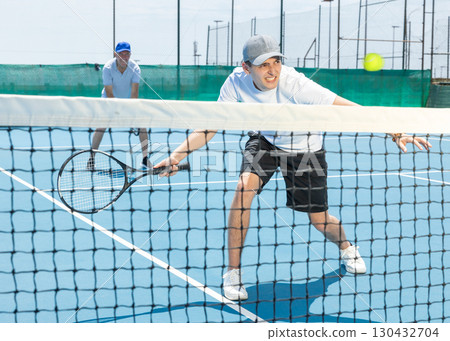 Portrait of young guy playing tennis on open court in summer, swinging racket to return ball over net. Sportsman ready to hit volley 130432704