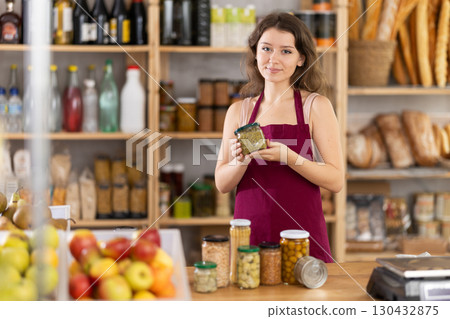 Young saleswoman is offering canned products in grocery store 130432875