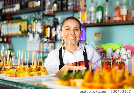 Friendly young woman bartender offering appetizing pinchos in pub Friendly young woman bartender offering appetizing pinchos in pub 130433107