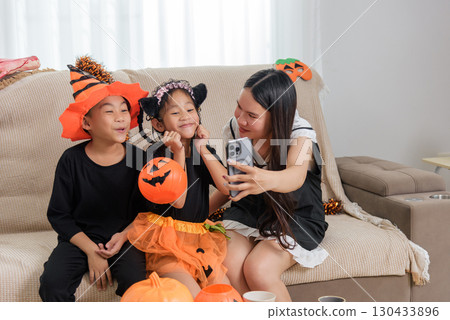 A woman is taking a photo with her phone of two children in costumes, a boy wearing an orange hat and a girl with cat ears holding a pumpkin bucket, they are all smiling on a sofa for halloween day. 130433896