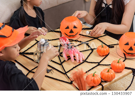 Children enjoy a Halloween celebration, preparing spooky crafts with pumpkins, spiders, and a fake severed hand on a tabletop covered in a spiderweb design and pipe cleaner details. 130433907