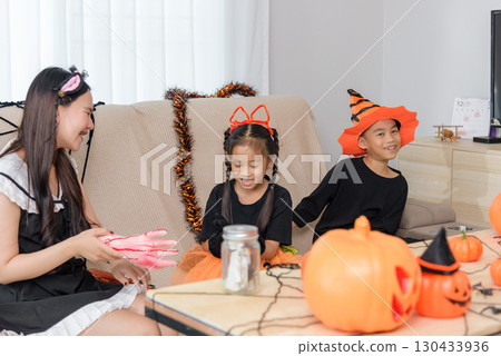 Smiling kids wear costumes, a woman is painting a fake arm with fake blood, all celebrating with pumpkins and garlands, sharing candy during festive halloween day preparations. 130433936